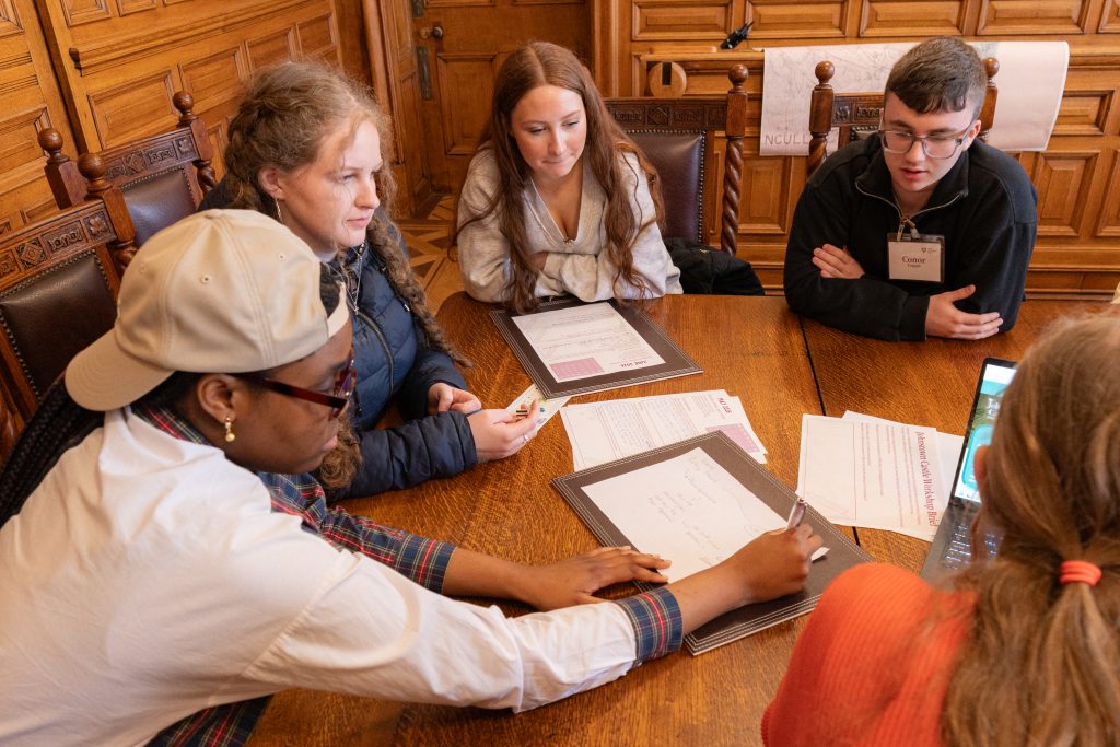 Five young people sitting around a table planning and writing using paper and a laptop