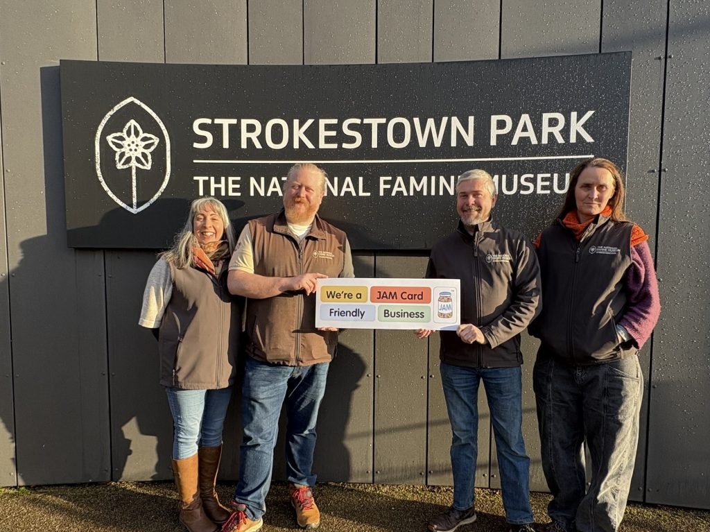 Two men hold a board which says 'We're a JAM Card Friendly Business' with two women either side of them. In the background is a large sign with "Strokestown Park,The National Famine Museum' and the logo of the potato flower on the left side.
