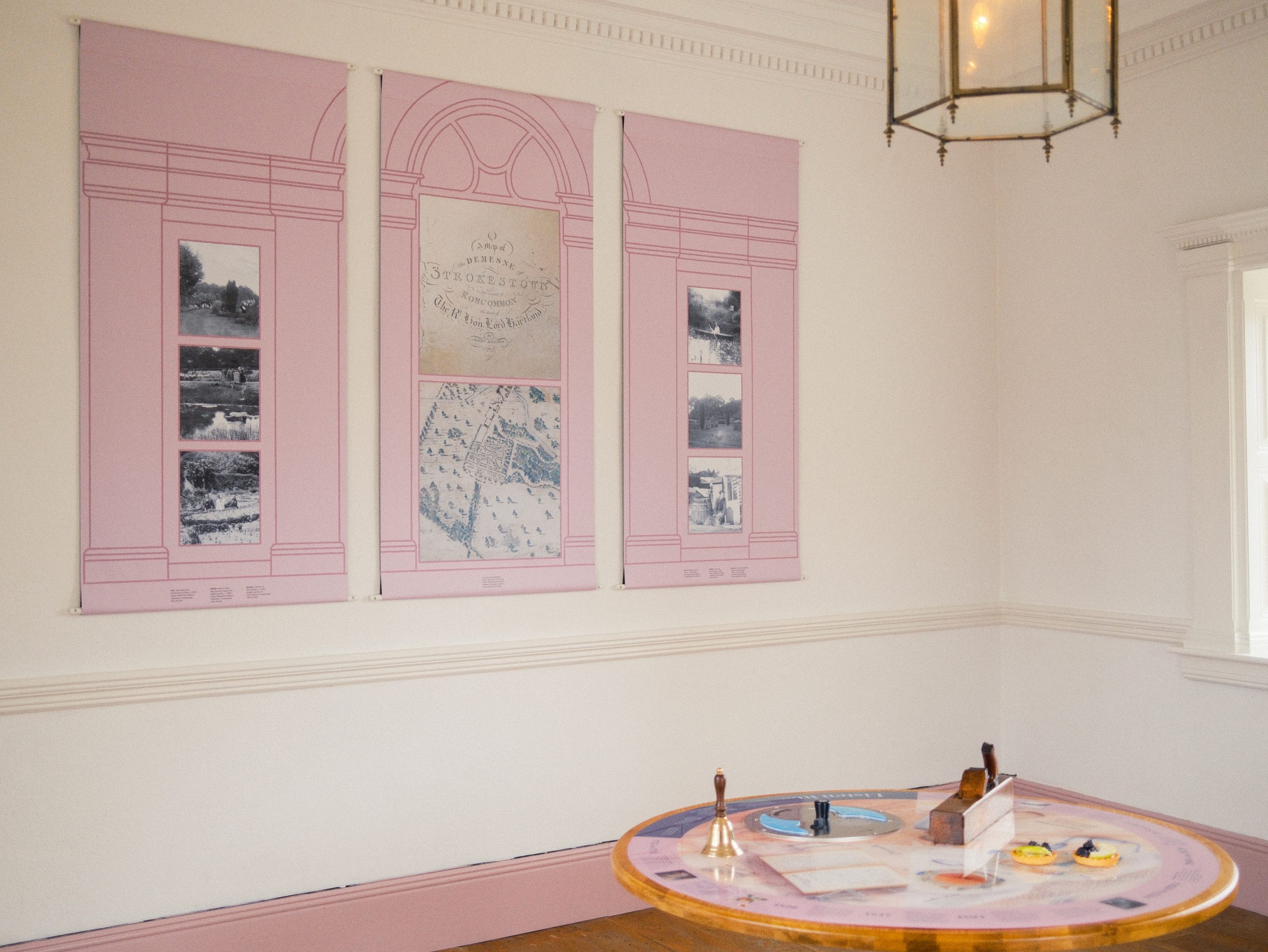To the foreground is a with a round wooden table with items detailing the story of the Gazebo and in the background there are three pink panels replicating windows and featuring photos of Strokestown Park over the centuries