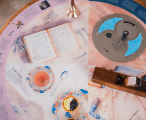A pink table features replicas and images of items telling the stories of those who frequented the Gazebo tower throughout the years. Including replica pastries, labourer tools and a bell sitting on the table and a background of text to follow along.
