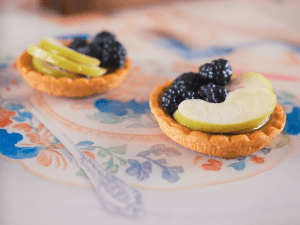 A pink table features replicas and images of items telling the stories of those who frequented the Gazebo tower throughout the years. Including replica pastries with glazed apple slices and blackberries sitting on the table and a background of text to follow along.