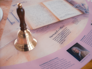 A pink table features replicas and images of items telling the stories of those who frequented the Gazebo tower throughout the years. Including a bell sitting on the table and a background of text to follow along.