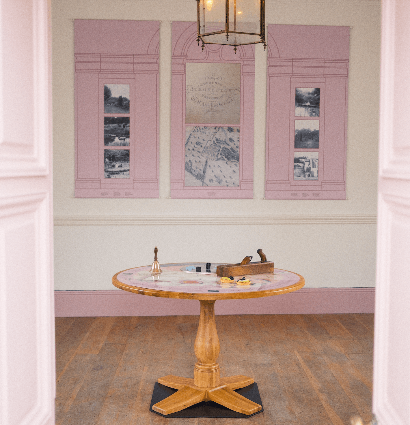 Open pink doors frame the image. Inside the room, to the foreground is a with a round wooden table with items detailing the story of the Gazebo and in the background there are three pink panels replicating windows and featuring photos of Strokestown Park over the centuries