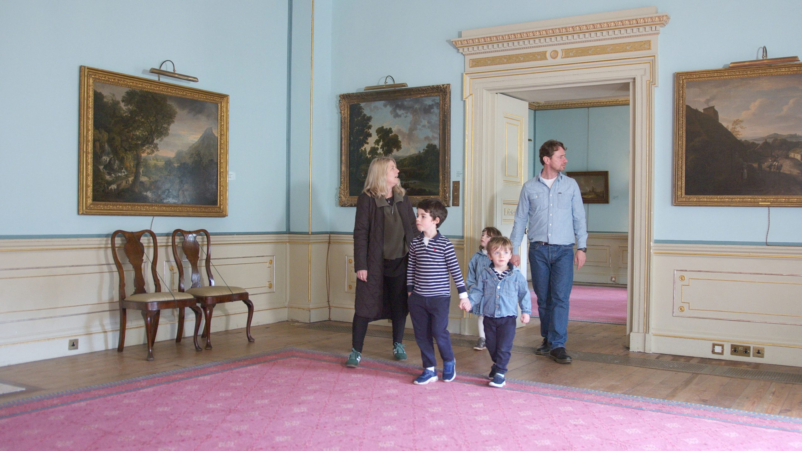 A woman, man and 3 children walk into a grand room in a Regency House with pale blue walls and period arts hanging on the walls.