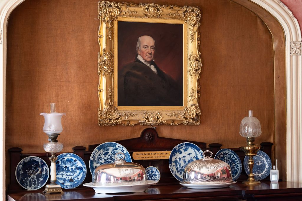A sideboard with the name George Baron Mount Sandford engraved on a gold plaque. There are blue and white period dishes and a portrait painting hanging above with a gold frame.