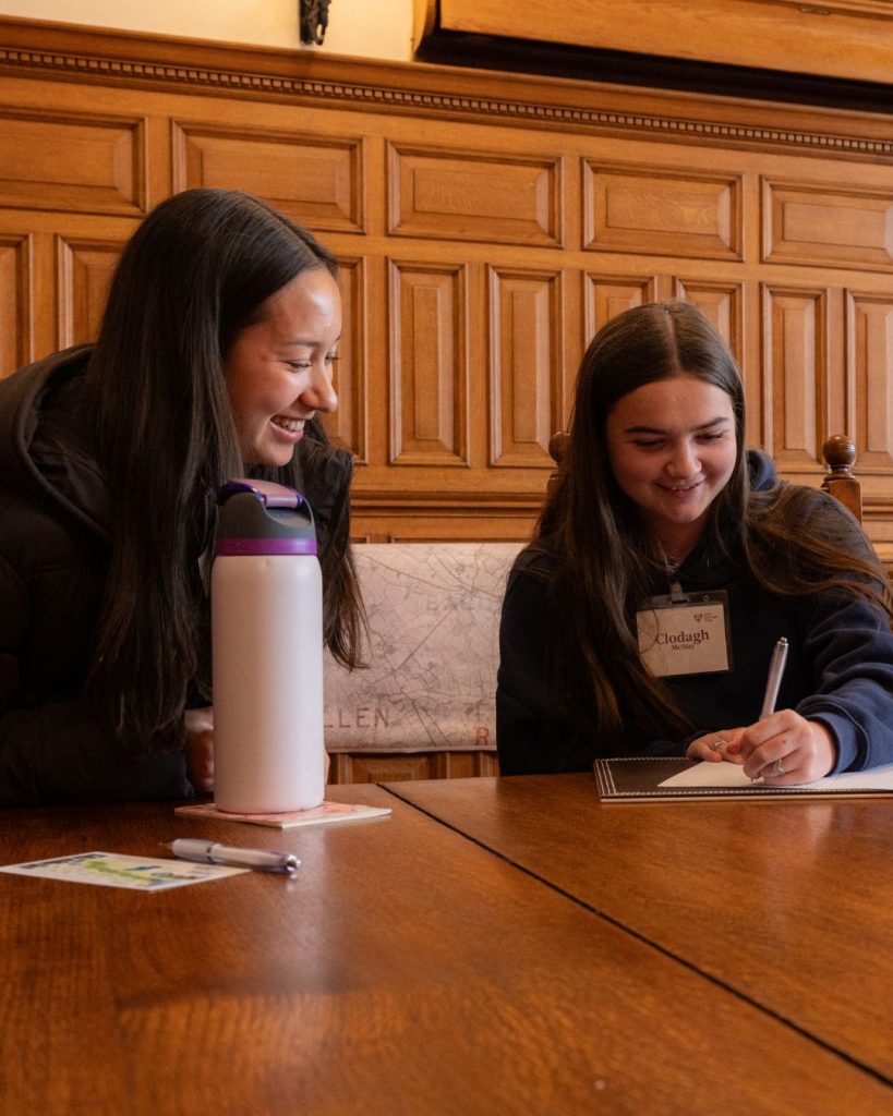 two transition year aged students smiling sitting at a large wooden table with paper in front of them