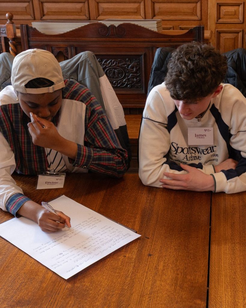 two transition year aged students smiling sitting at a large wooden table with paper in front of them