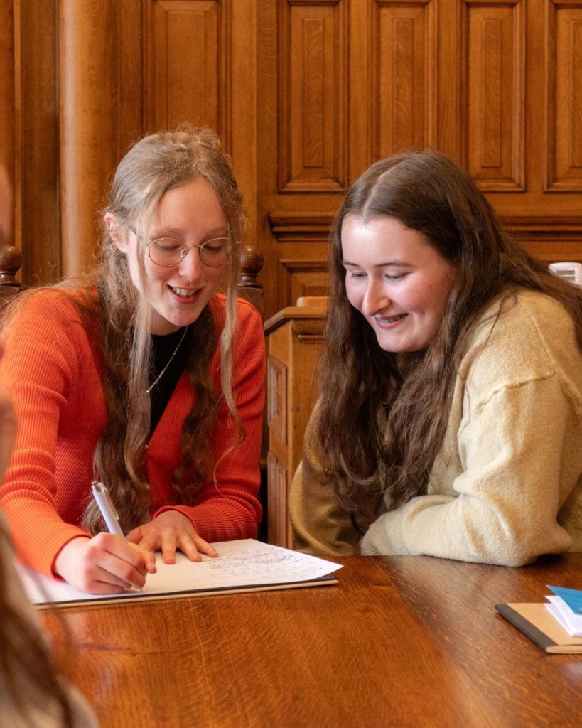 two transition year aged students smiling sitting at a large wooden table with paper in front of them
