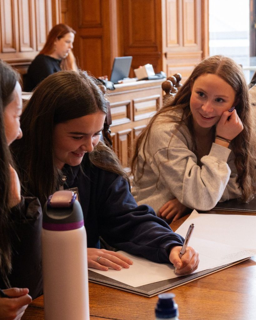 two transition year aged students smiling sitting at a large wooden table with paper in front of them
